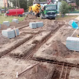 Przygotowanie fundamentów pod budowę domu: wykopy, zbrojenie, palety z bloczkami betonowymi, niwelator laserowy na trójnogu, betoniarka i ciężarówka na działce budowlanej.