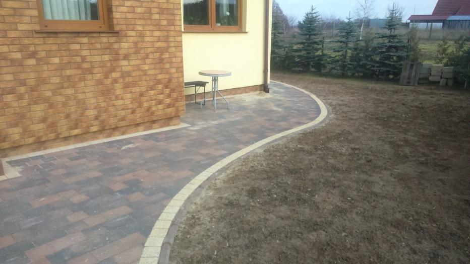 Ułożona ścieżka z kostki brukowej w odcieniach brązu i szarości, obrzeżona jasną kostką, prowadząca od ściany budynku z cegły klinkierowej do trawnika; w tle widoczne iglaki.