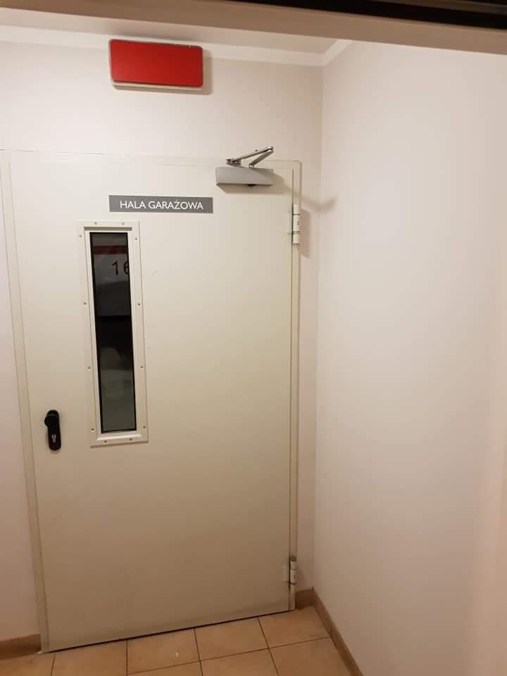 Białe drzwi do hali garażowej z małym okienkiem i napisem 'Hala Garażowa' nad nim, zamontowane w białej ścianie, z widocznym automatem samozamykającym.