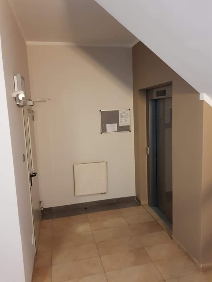 Korytarz po remoncie z płytkami podłogowymi w odcieniach beżu, świeżo pomalowane ściany w kolorze kremowym i beżowym, grzejnik na ścianie i metalowa winda w tle.