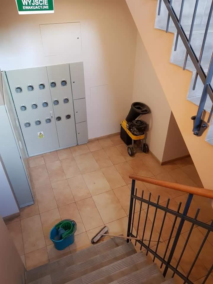 Klatka schodowa z terakotą po umyciu, widoczne wiadro z mopem, szczotka, osuszacz powietrza i szafa elektryczna z tabliczką ostrzegawczą.