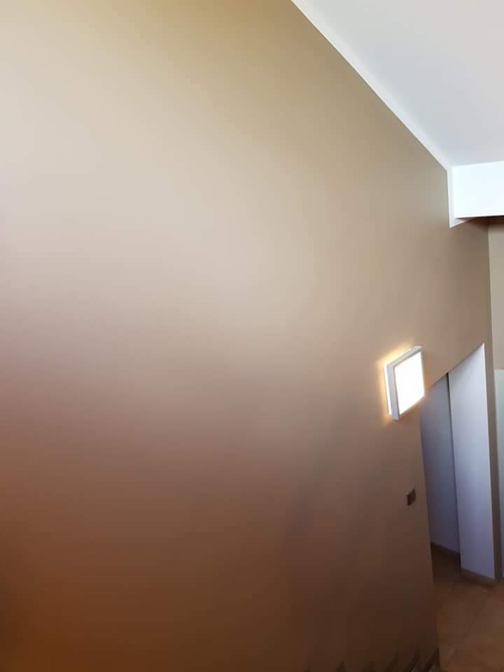 Ściana w odcieniach beżu po świeżym malowaniu, z minimalistycznym oświetleniem ściennym i widocznym fragmentem sufitu.