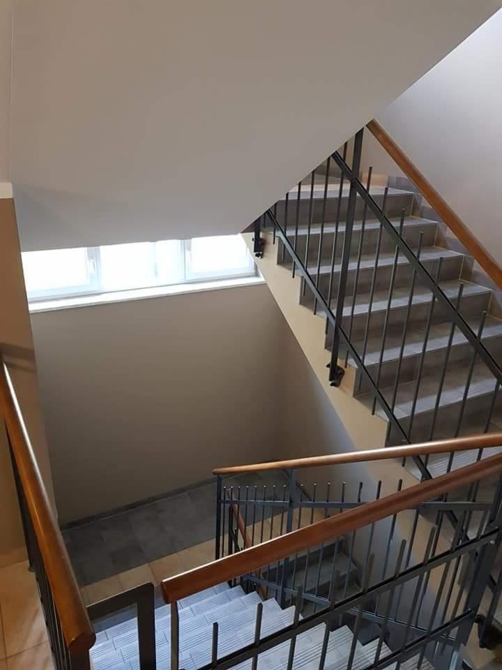 Klatka schodowa z metalową balustradą i drewnianymi poręczami, widok z góry, ściany pomalowane na beżowo, okno na półpiętrze.