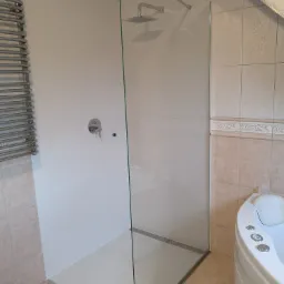 Nowoczesna kabina prysznicowa ze szklaną ścianą w łazience z beżowymi płytkami, widoczny panel sterowania prysznicem i grzejnik drabinkowy.