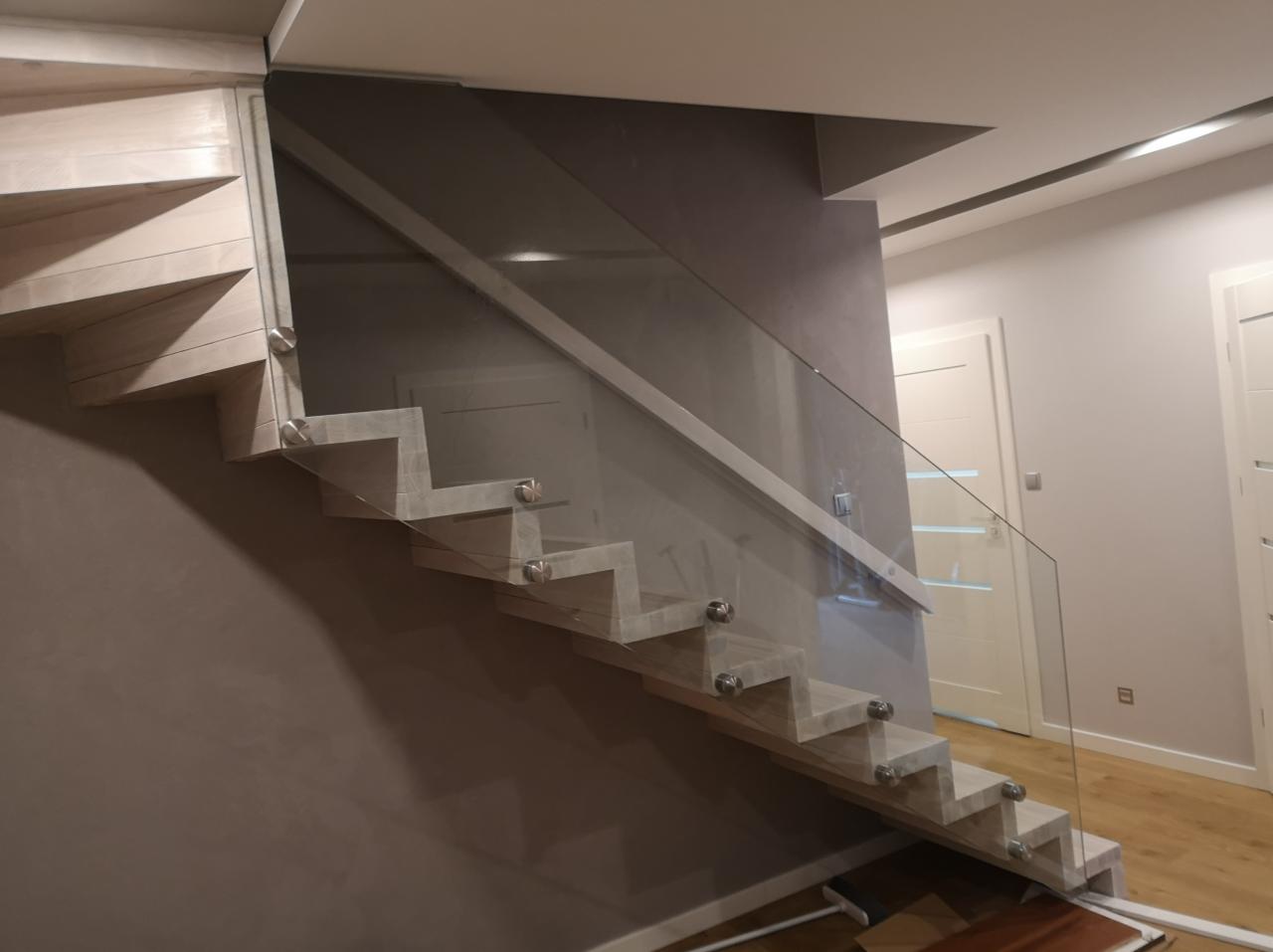 Nowoczesne schody z drewnianymi stopniami i szklaną balustradą, mocowaną za pomocą metalowych uchwytów punktowych, na tle ściany w odcieniach szarości.