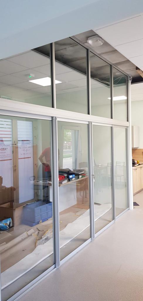 Szklana ścianka działowa z przesuwnymi drzwiami w biurze, widoczne niedokończone prace wykończeniowe, aluminiowe profile, widok na pomieszczenie biurowe z wyposażeniem.