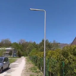 Nowoczesna srebrna latarnia uliczna z pojedynczym ramieniem oświetlająca żwirową ścieżkę wzdłuż ogrodzenia z żywopłotu, błękitne niebo w tle.