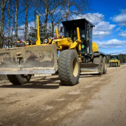 Żółty równiarka drogowa z brudną lemieszą na polnej drodze, w tle drzewa i błękitne niebo z chmurami, w oddali widoczny walec drogowy.