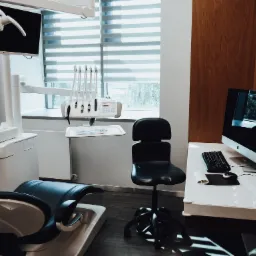 Nowoczesny gabinet stomatologiczny z fotelem, lampą zabiegową, narzędziami, komputerem iMac i roletami okiennymi.