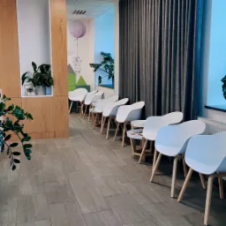 Jasne, przestronne wnętrze poczekalni z rzędem białych krzeseł na drewnianych nogach, roślinami doniczkowymi i drewnianą ścianą z wnęką.