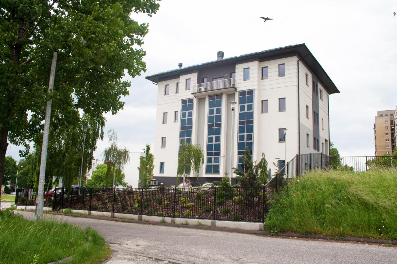 Nowoczesny budynek z balkonem i dużymi oknami, otoczony zielenią i ogrodzeniem, widziany z poziomu ulicy.