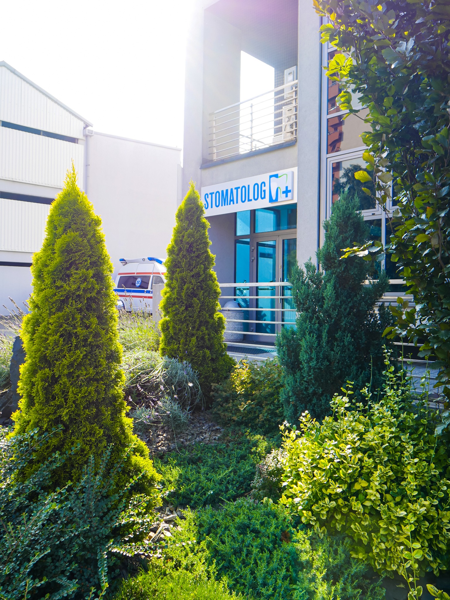Budynek gabinetu z szyldem i logo, zieleń ozdobna przed wejściem, w tle zaparkowany ambulans.