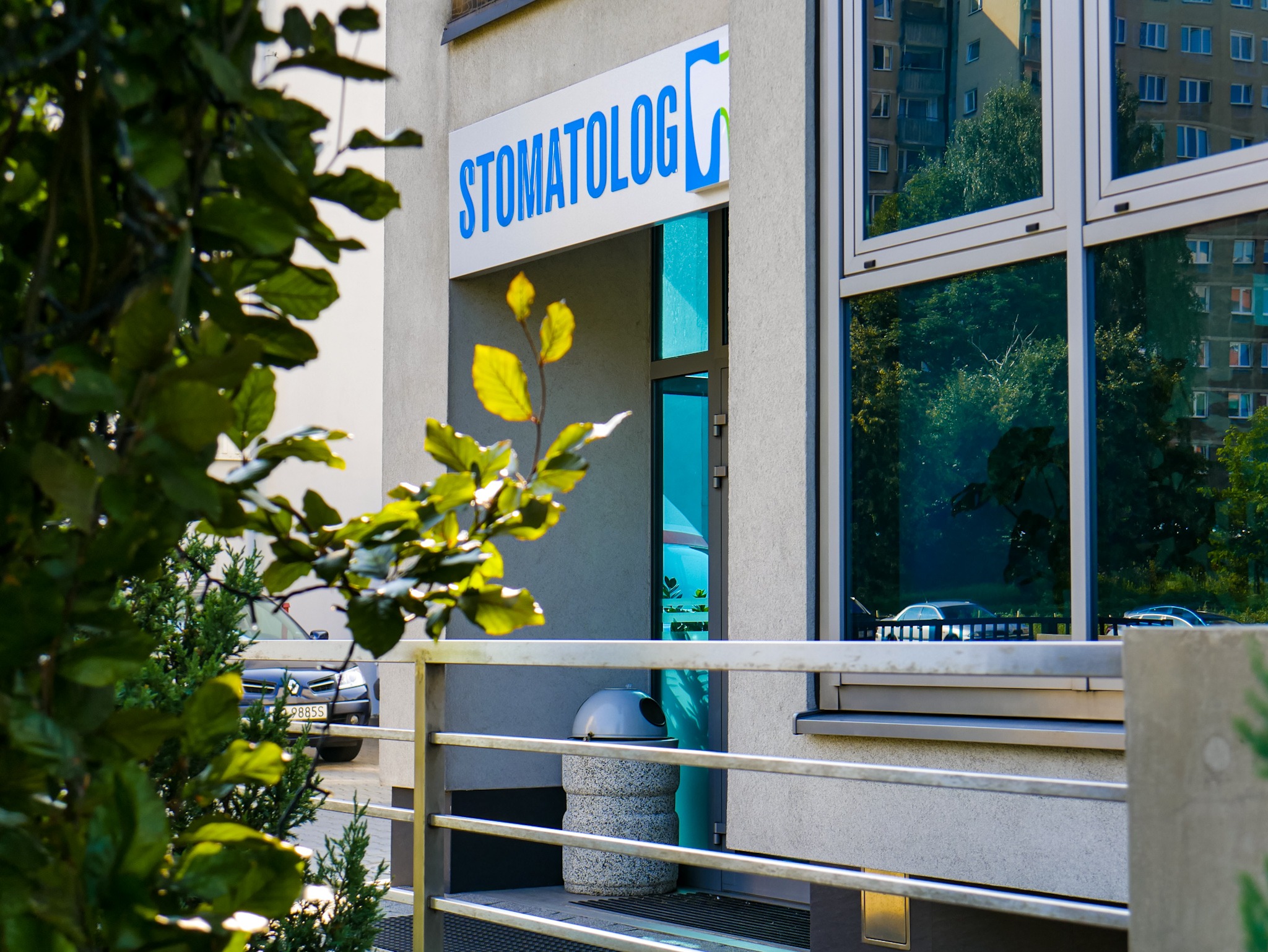 Wejście do budynku z niebieskim szyldem z napisem w języku polskim, za metalową barierką, częściowo zasłonięte zielenią drzew.