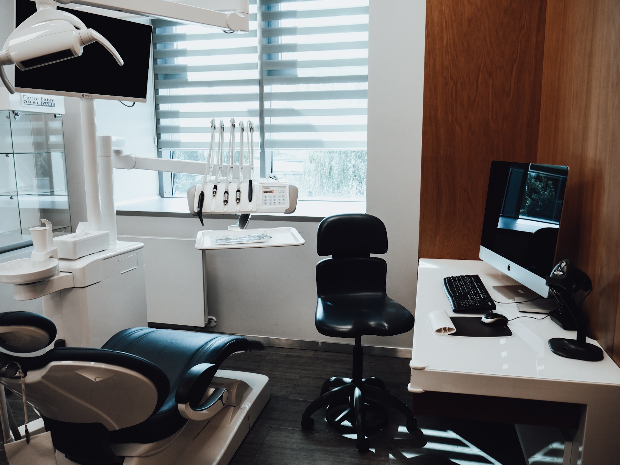 Nowoczesny gabinet stomatologiczny z fotelem, lampą zabiegową, narzędziami, komputerem iMac i roletami okiennymi.
