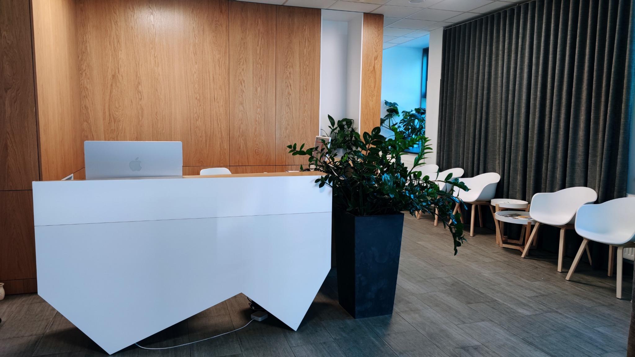 Jasne wnętrze poczekalni z białą recepcją, laptopem Apple, rośliną w czarnej donicy i rzędem białych krzeseł na drewnianych nogach pod ciemnymi zasłonami.