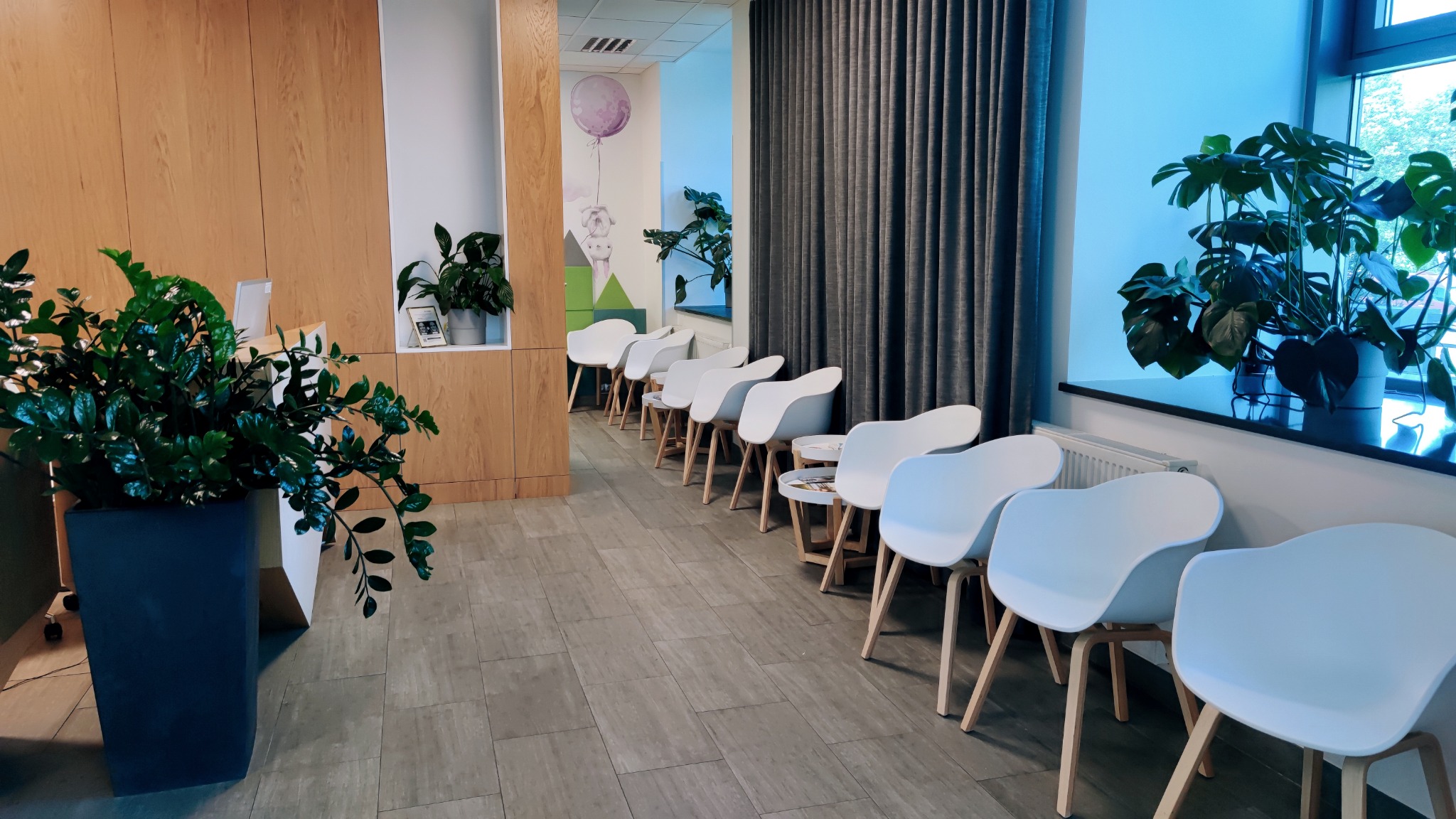 Jasne, przestronne wnętrze poczekalni z rzędem białych krzeseł na drewnianych nogach, roślinami doniczkowymi i drewnianą ścianą z wnęką.