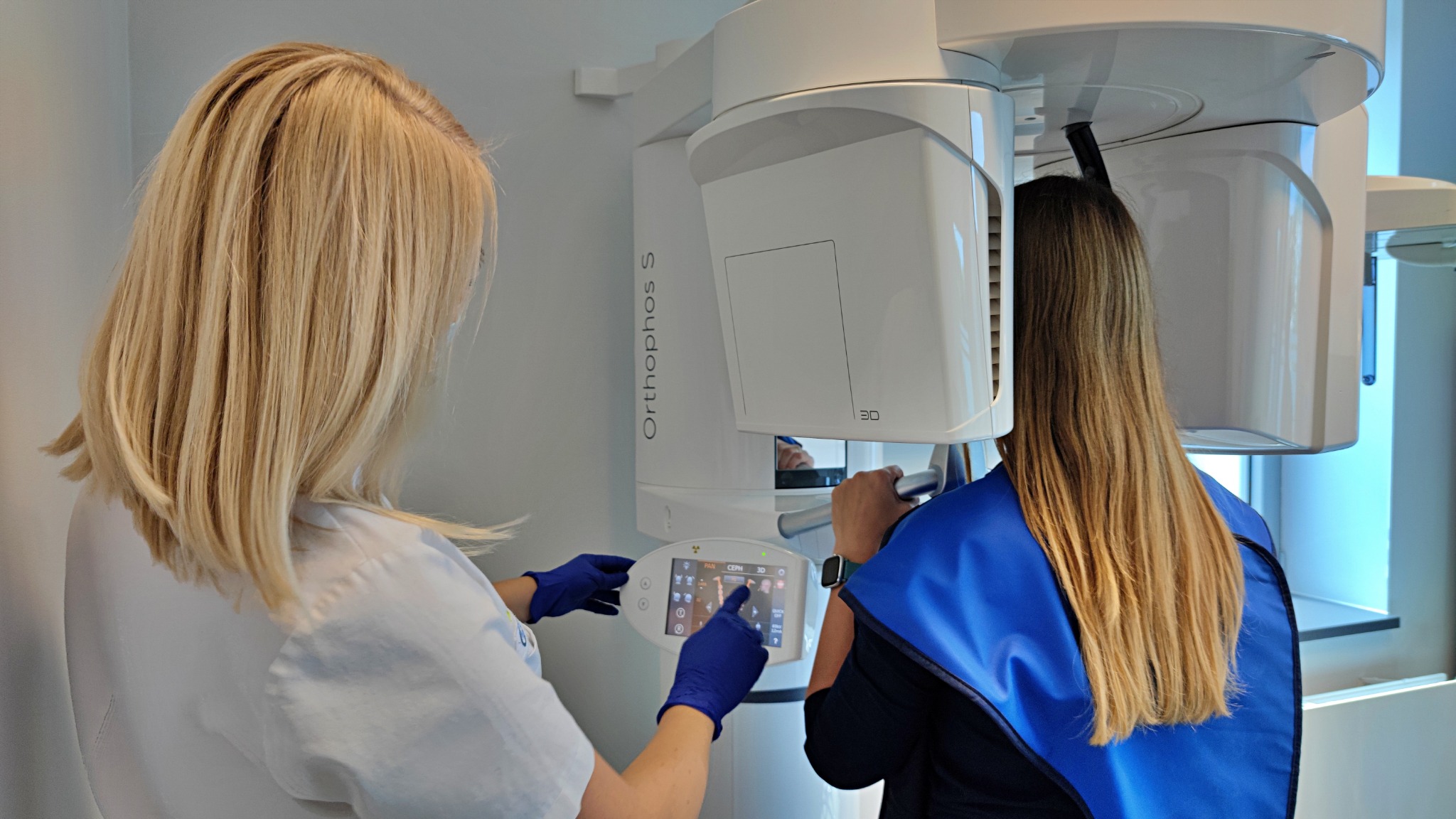 Pacjentka podczas badania pantomograficznego w gabinecie, obsługa urządzenia przez osobę w uniformie medycznym i niebieskich rękawiczkach, widoczny panel dotykowy z ikonami.