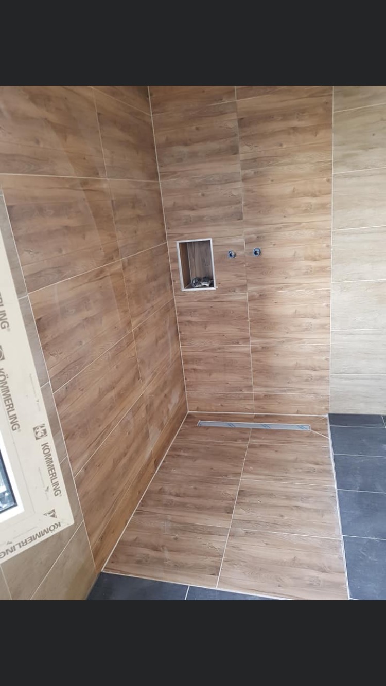 Kabina prysznicowa wyłożona brązowymi płytkami imitującymi drewno, z odpływem liniowym i wnęką na kosmetyki, widoczne dwa przyłącza wodne, fragment okna z profilem Kommerling.