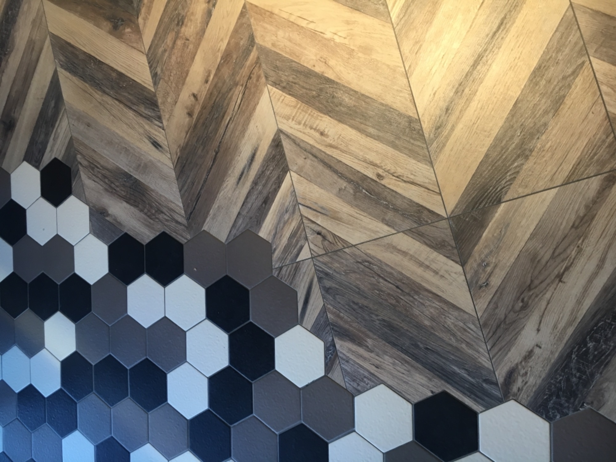 Fragment ściany z dekoracyjnym połączeniem płytek drewnopodobnych ułożonych w jodełkę i heksagonalnych płytek w odcieniach szarości, bieli i czerni.