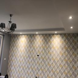 Darex - Pokój po remoncie z tapetą w geometryczne wzory, nowoczesny żyrandol z czarnymi abażurami i oświetlenie sufitowe punktowe.