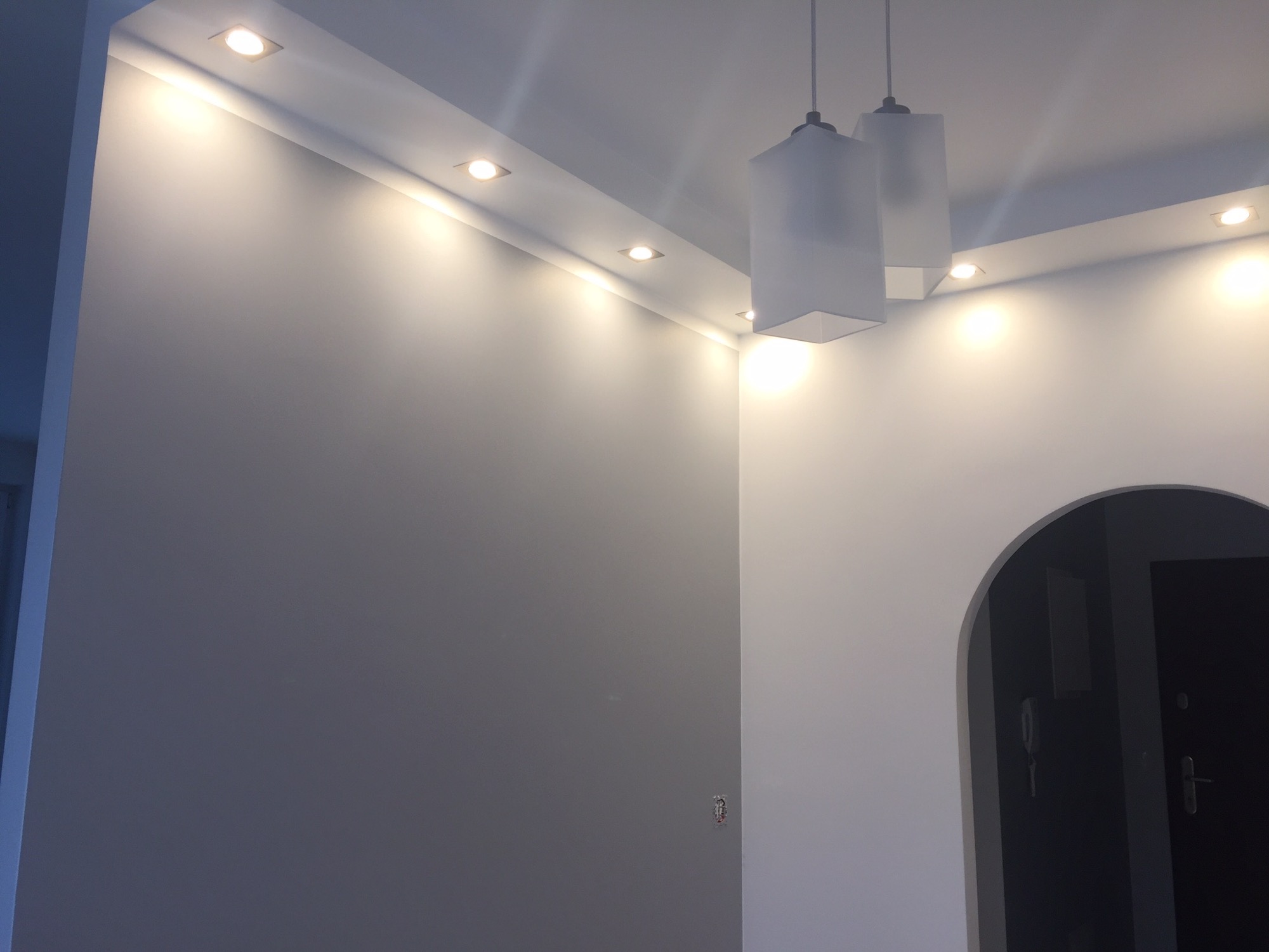 Szara ściana z oświetleniem punktowym LED i dwoma wiszącymi lampami z białym abażurem w nowocześnie wykończonym wnętrzu z łukowatym przejściem do innego pomieszczenia.