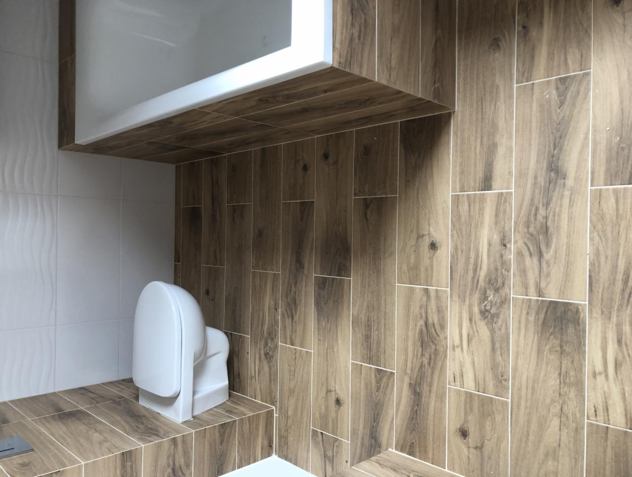 Nowoczesna łazienka z toaletą bezkołnierzową, brązowe płytki imitujące drewno na ścianach i podłodze, biała zabudowa nad toaletą, białe płytki z delikatną fakturą w tle.