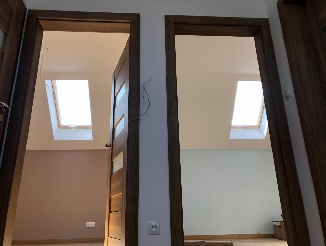 Dwa drewniane ościeżnice z widokiem na poddasze z oknami dachowymi, ściany pomalowane na różne kolory, widoczne gniazdka elektryczne i wystające kable.