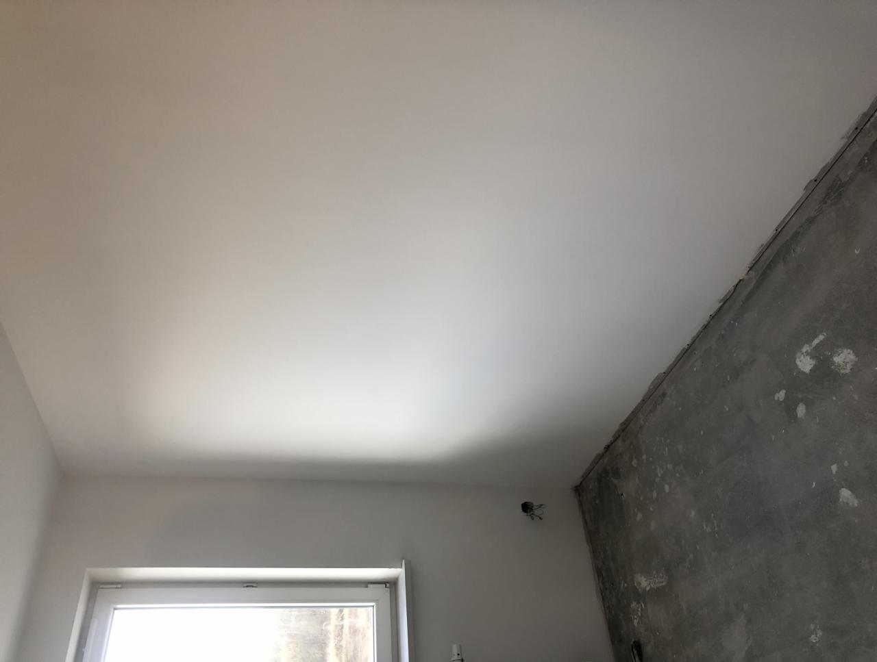 Wnętrze pokoju w trakcie prac wykończeniowych w Krakowie: gładki, pomalowany sufit kontrastuje z surową, betonową ścianą. Widoczne okno i wystające przewody elektryczne.