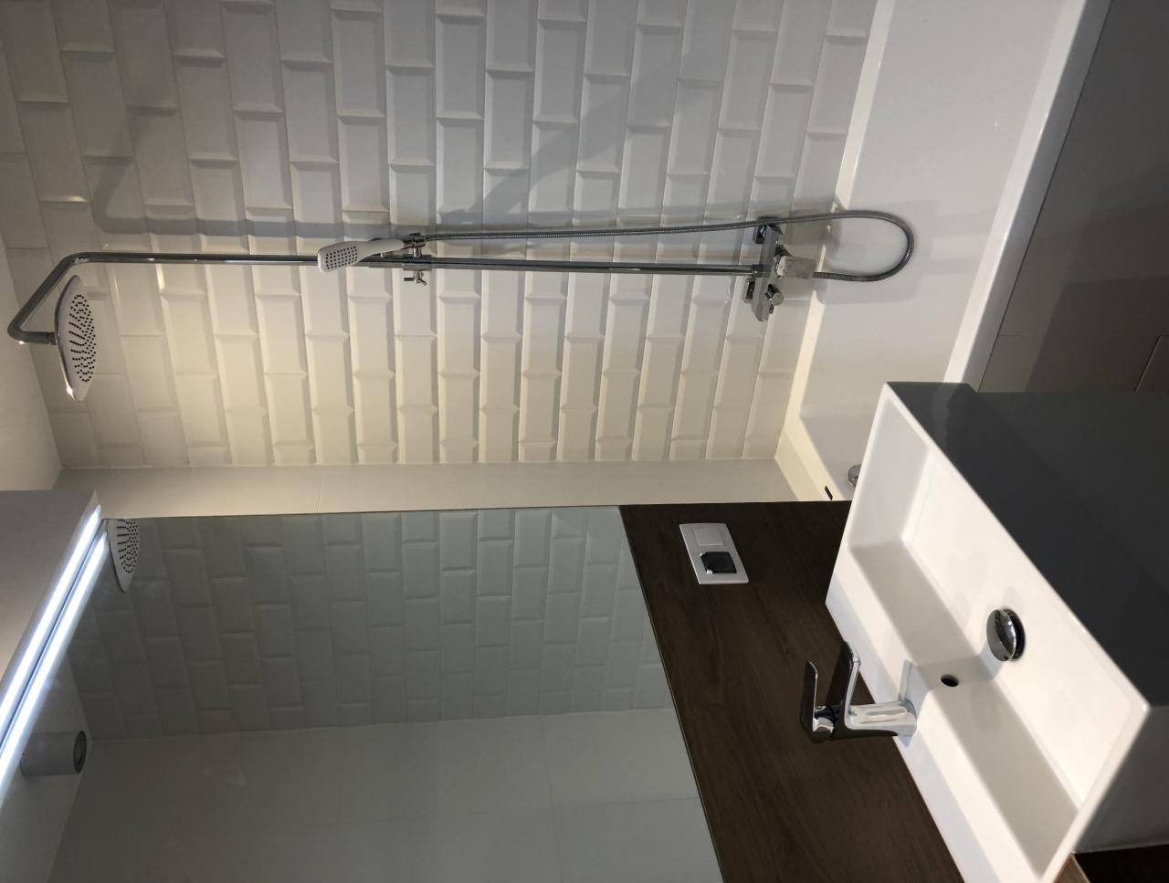 Nowoczesna łazienka z białymi, prostokątnymi płytkami na ścianie, chromowaną baterią prysznicową z deszczownicą i słuchawką oraz prostokątną umywalką nablatową na blacie z ciemnego drewna.