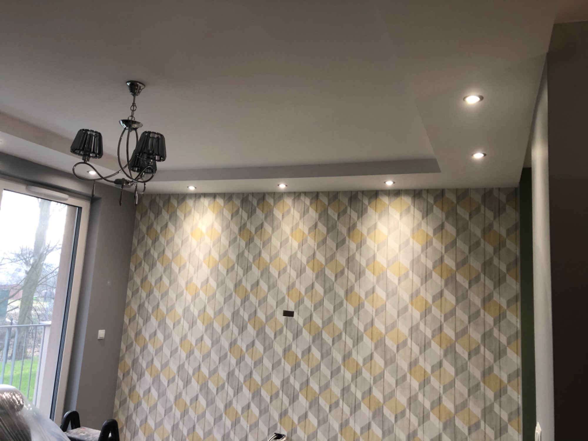 Pokój po remoncie z tapetą w geometryczne wzory, nowoczesny żyrandol z czarnymi abażurami i oświetlenie sufitowe punktowe.