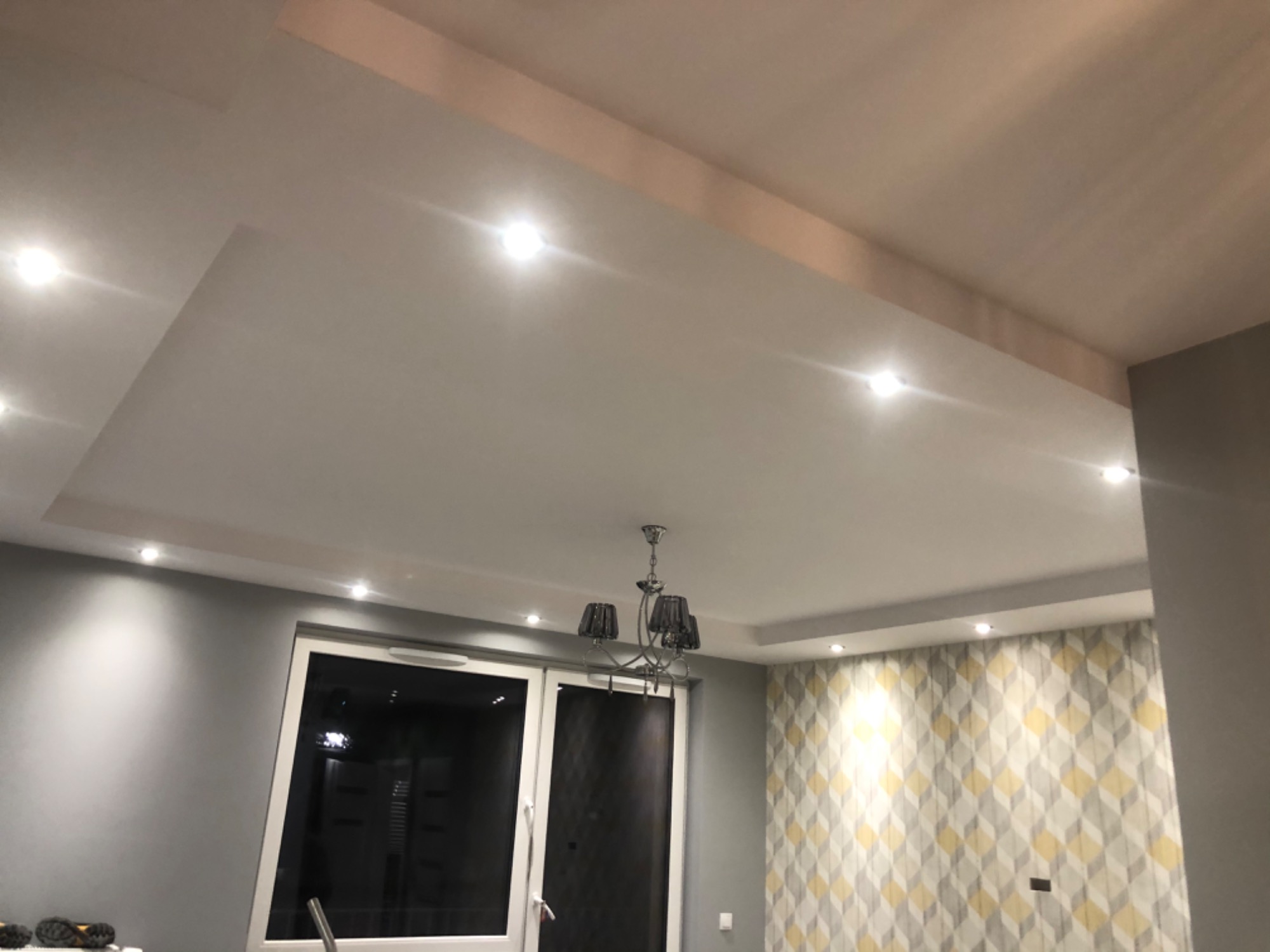 Sufit podwieszany z oświetleniem punktowym i ozdobnym żyrandolem w pokoju z szarą ścianą i tapetą w geometryczne wzory.