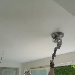 Szlifowanie sufitu za pomocą szlifierki z odkurzaczem, widoczne okno z zasłoniętą folią szybą, fragment ściany i sufitu po obróbce.