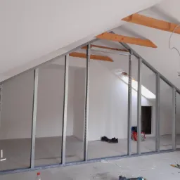 Konstrukcja szkieletowa z profili metalowych na poddaszu, przygotowana do montażu płyt gipsowo-kartonowych. Widoczne drewniane belki stropowe, okno dachowe, narzędzia i materiały budowlane...