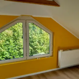 Pokój na poddaszu z żółtymi ścianami, trójkątnym oknem z widokiem na zieleń i białym grzejnikiem panelowym.