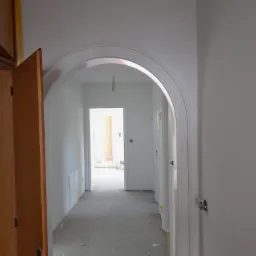 Korytarz w trakcie remontu, ściany świeżo pomalowane na biało, widoczne ślady farby na podłodze, otwarte drzwi po lewej stronie, łukowe przejście do dalszej części korytarza.