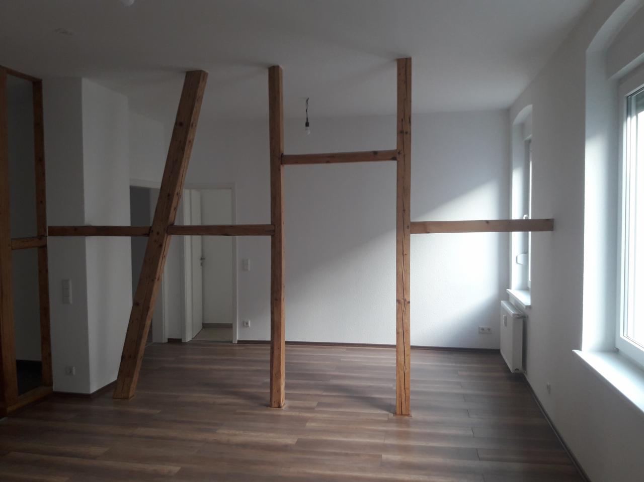 Wnętrze w trakcie przebudowy z widoczną drewnianą konstrukcją wsporczą, jasnymi ścianami, panelami podłogowymi w odcieniach brązu i białym grzejnikiem pod oknem.
