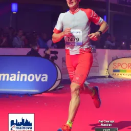 Biegacz Piotr Tomczyk w czerwono-białym stroju, z numerem 7479, biegnie podczas Mainova Frankfurt Marathon, z czasem 2:44:10, widoczny fragment mety i kibiców.