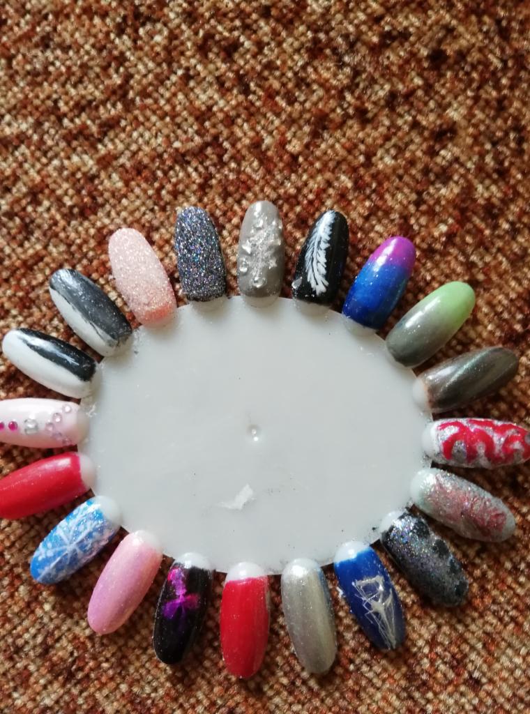 Wzornik z różnorodnymi stylizacjami paznokci, prezentujący zdobienia z brokatem, efektem ombre, motywem śnieżynki, piórka i innymi detalami na plastikowym kole na tle brązowej tkaniny.