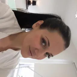 Uśmiechnięta kobieta w białym uniformie prezentuje przedłużone rzęsy, z widocznym subtelnym makijażem i oświetleniem lampą kosmetyczną w tle.