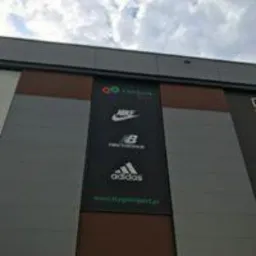 Wielkoformatowy baner reklamowy z logo marek sportowych (Olympus, Nike, New Balance, Adidas) umieszczony na fasadzie budynku.