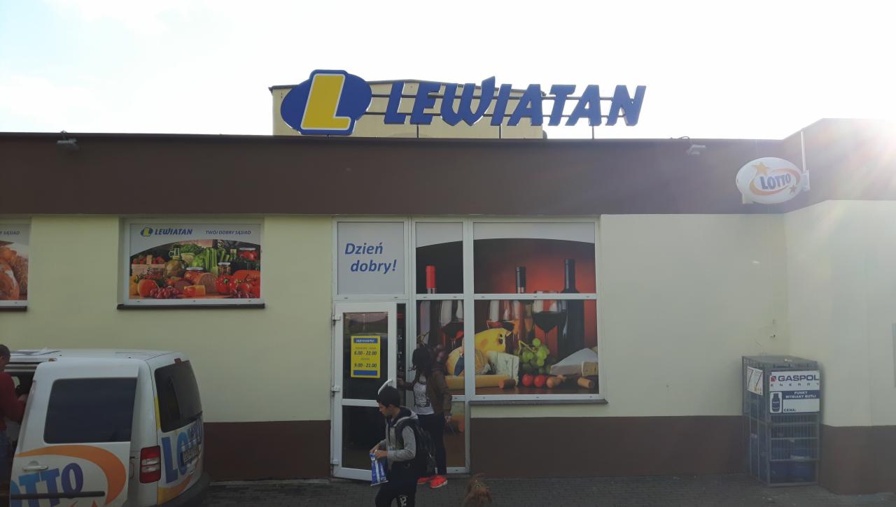 Fasada sklepu spożywczego z szyldem, witrynami oklejonymi grafikami przedstawiającymi produkty spożywcze, oraz oklejonym samochodem dostawczym w tle.