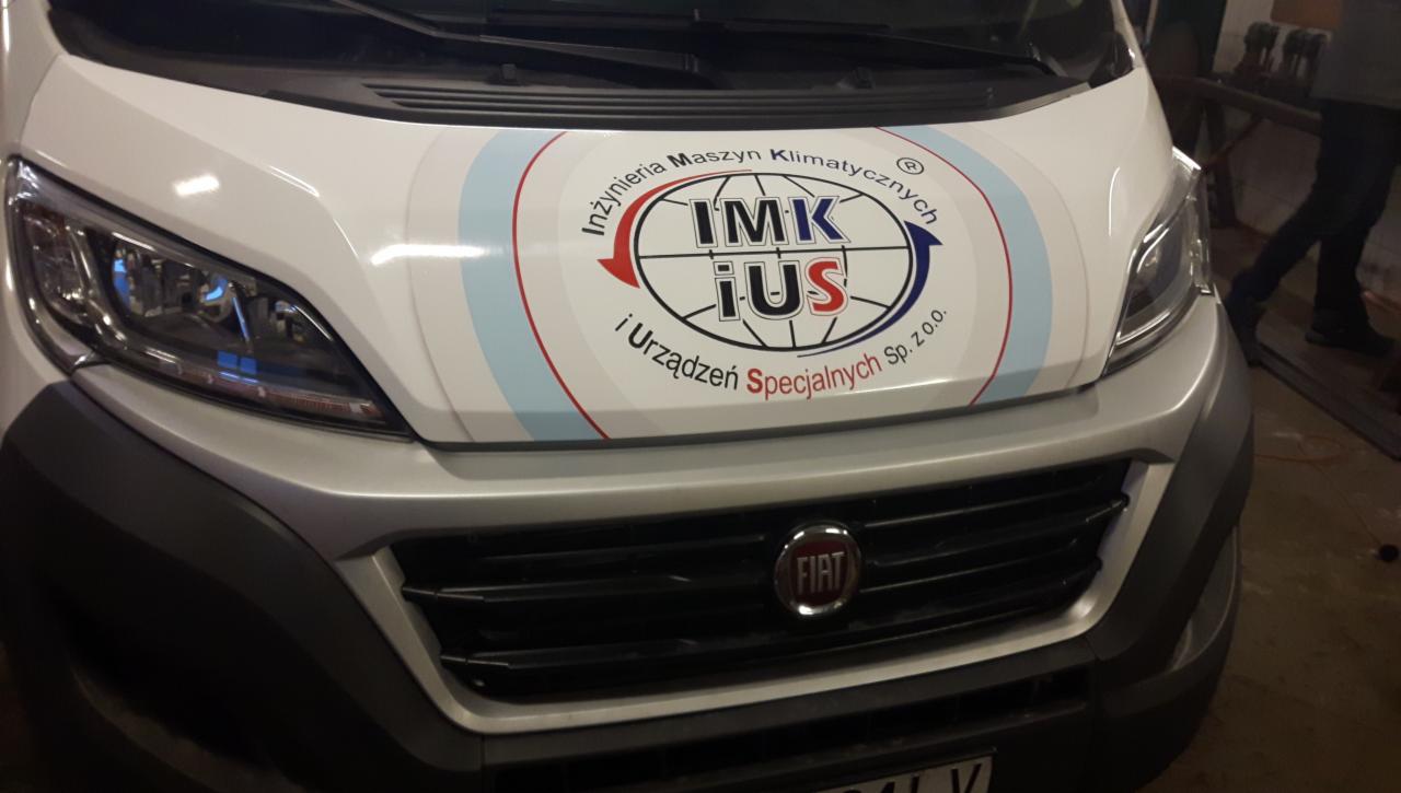 Srebrny samochód dostawczy Fiat z naklejoną grafiką firmową na masce przedstawiającą logo firmy 'IMK iUS' w otoczeniu napisu 'Inżynieria Maszyn Klimatycznych' i 'i Urządzeń Specjalnych Sp. z o.o.'