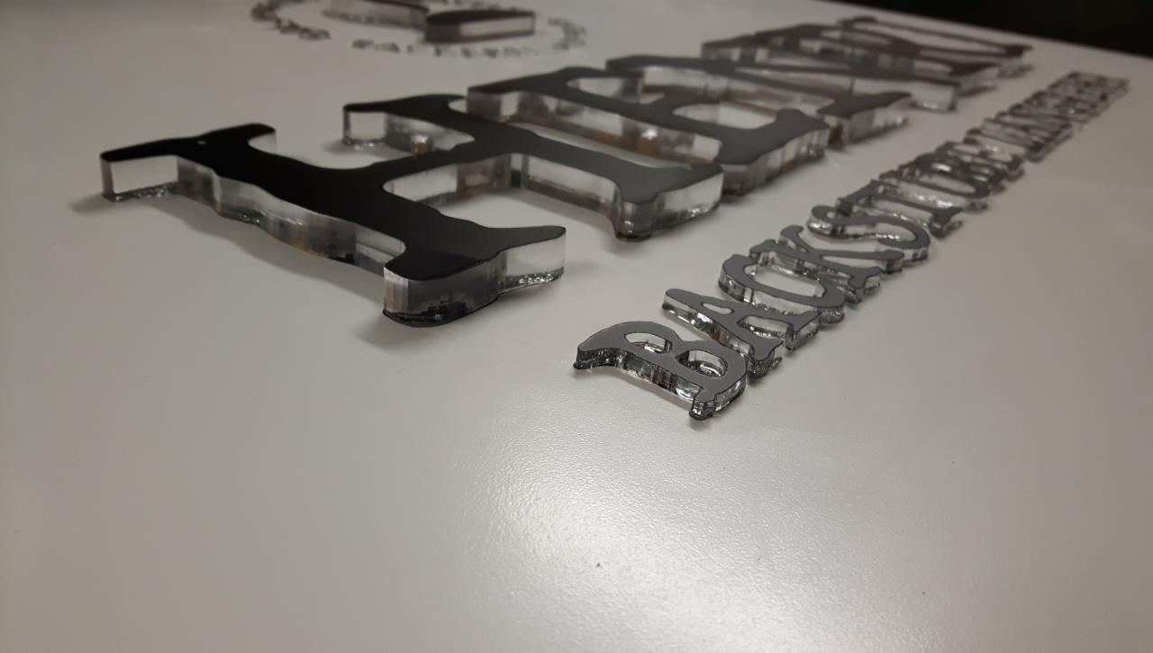 Perspektywiczne ujęcie akrylowych liter tworzących napisy, częściowo czarne, częściowo srebrne, z efektem trójwymiarowości, leżące na białym podłożu.