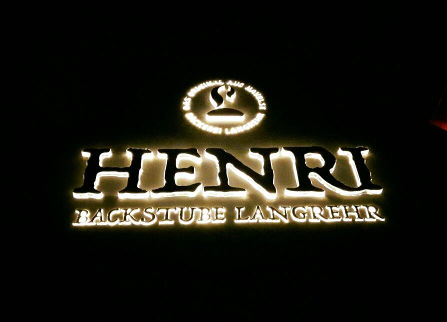 Podświetlany szyld z nazwą 'HENRI BACKSTUBE LANGREHR' oraz logo piekarni, zamontowany na ciemnej ścianie.