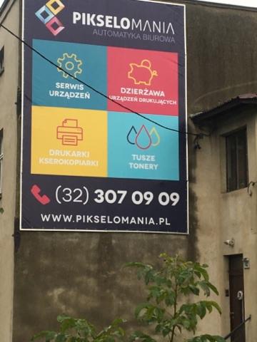 Duży baner reklamowy firmy Pikselomania umieszczony na ścianie budynku, oferujący serwis urządzeń, dzierżawę urządzeń drukujących, sprzedaż drukarek/kserokopiarek oraz tuszy/tonerów.