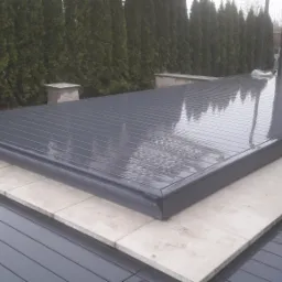 Szara, zadaszona konstrukcja tarasowa z automatycznym systemem zwijania, otoczona płytkami z jasnego kamienia i ciemnoszarą drewnianą podłogą, widok na rząd wysokich, zielonych tui w tle, lekki...