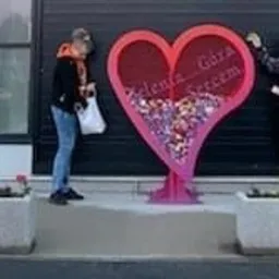 Dwie osoby umieszczają ozdoby w metalowej konstrukcji w kształcie serca z napisem, stojącej przed budynkiem z przeszkloną fasadą, w tle widoczne tuje w betonowych donicach.