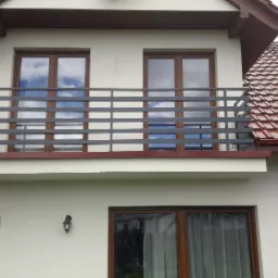 Dwukondygnacyjny dom z balkonem z szarą, poziomą balustradą, widok z dołu na dach z czerwonej dachówki i fragment złożonego parasola ogrodowego.