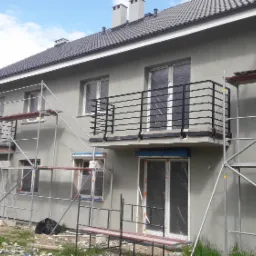 Nowe, szare szeregówki z czarnymi, prostymi balustradami balkonowymi, częściowo osłonięte rusztowaniami podczas prac wykończeniowych.