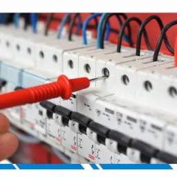 Pomiar napięcia w rozdzielnicy elektrycznej za pomocą próbnika napięcia z czerwoną rączką, widoczne przewody w kolorach niebieskim, czarnym i czerwonym.